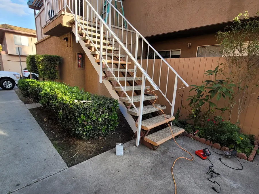 Exterior wood stairway with a white metal handrail at a stucco community building, with a hose and tools left on the concrete below