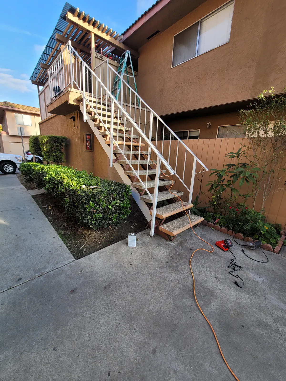 Exterior wood stairway with a white metal handrail running up to an upper unit at a stucco community building, with a hose and tools on the concrete below