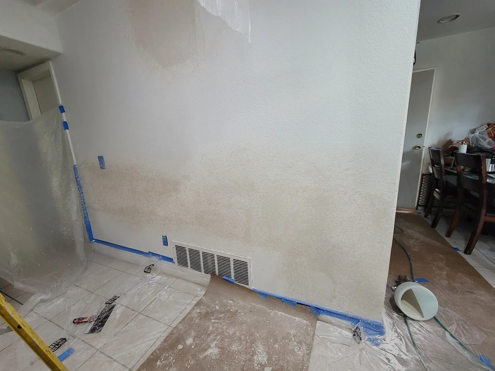 Interior wall masked at the baseboard and ceiling, with a large drywall patch feathered and textured to match the surrounding wall and taping tools on the covered floor