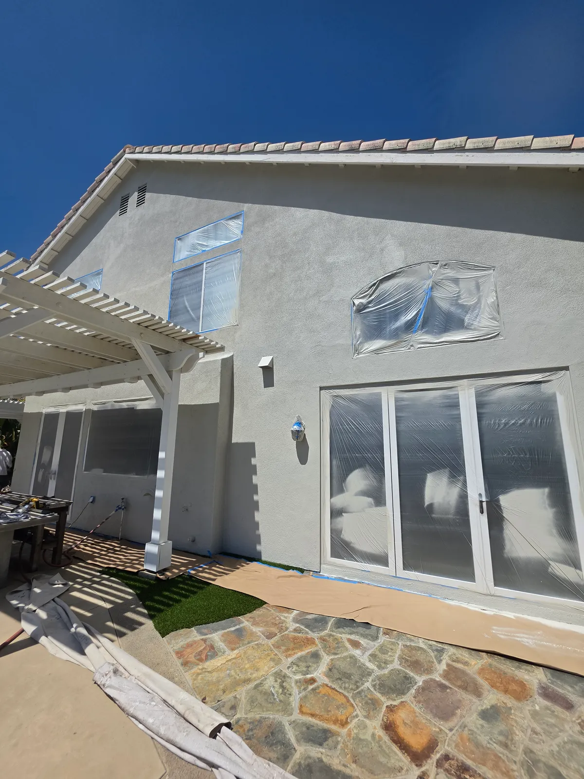 Two-story house with freshly painted light stucco, its windows and sliding doors covered in plastic sheeting and blue tape, and drop cloths laid over the flagstone patio