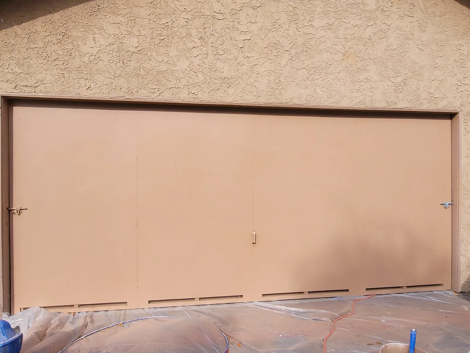 Wide garage door freshly repainted in a warm brown, set in a heavily textured stucco wall, with plastic sheeting still covering the driveway below