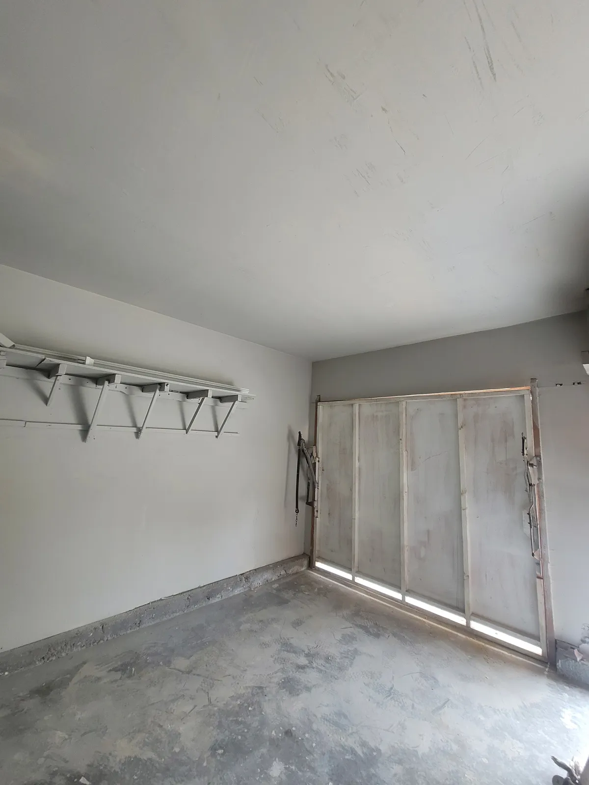 Inside of a garage with the walls and ceiling freshly painted white, a steel shelf bracket rail on the left and a paneled roll-up door on the right