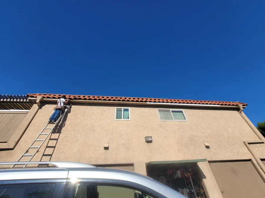 Extension ladder set against the gutter of a two story stucco building with a tile roof, one of the crew working at the roof line