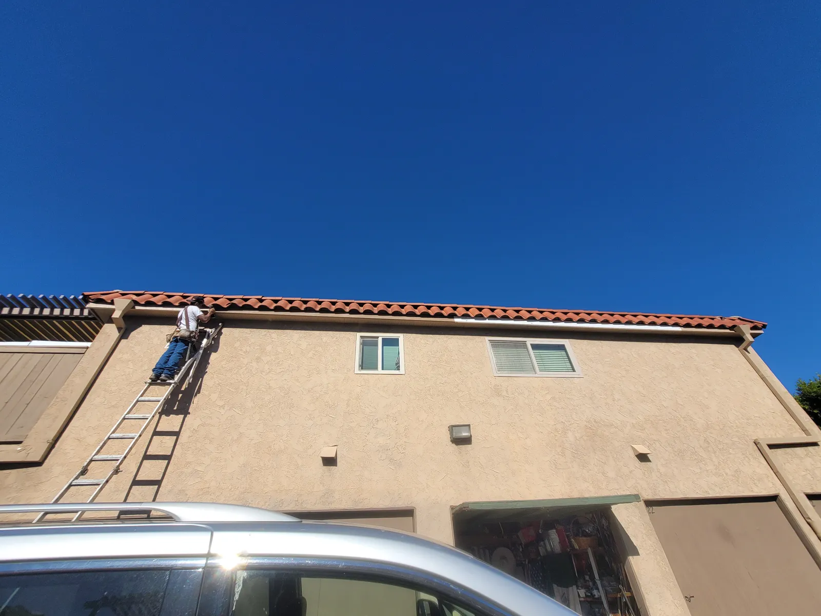 An extension ladder set against the gutter of a two-story stucco building with a tile roof, one of the crew working at the roof line under a clear blue sky