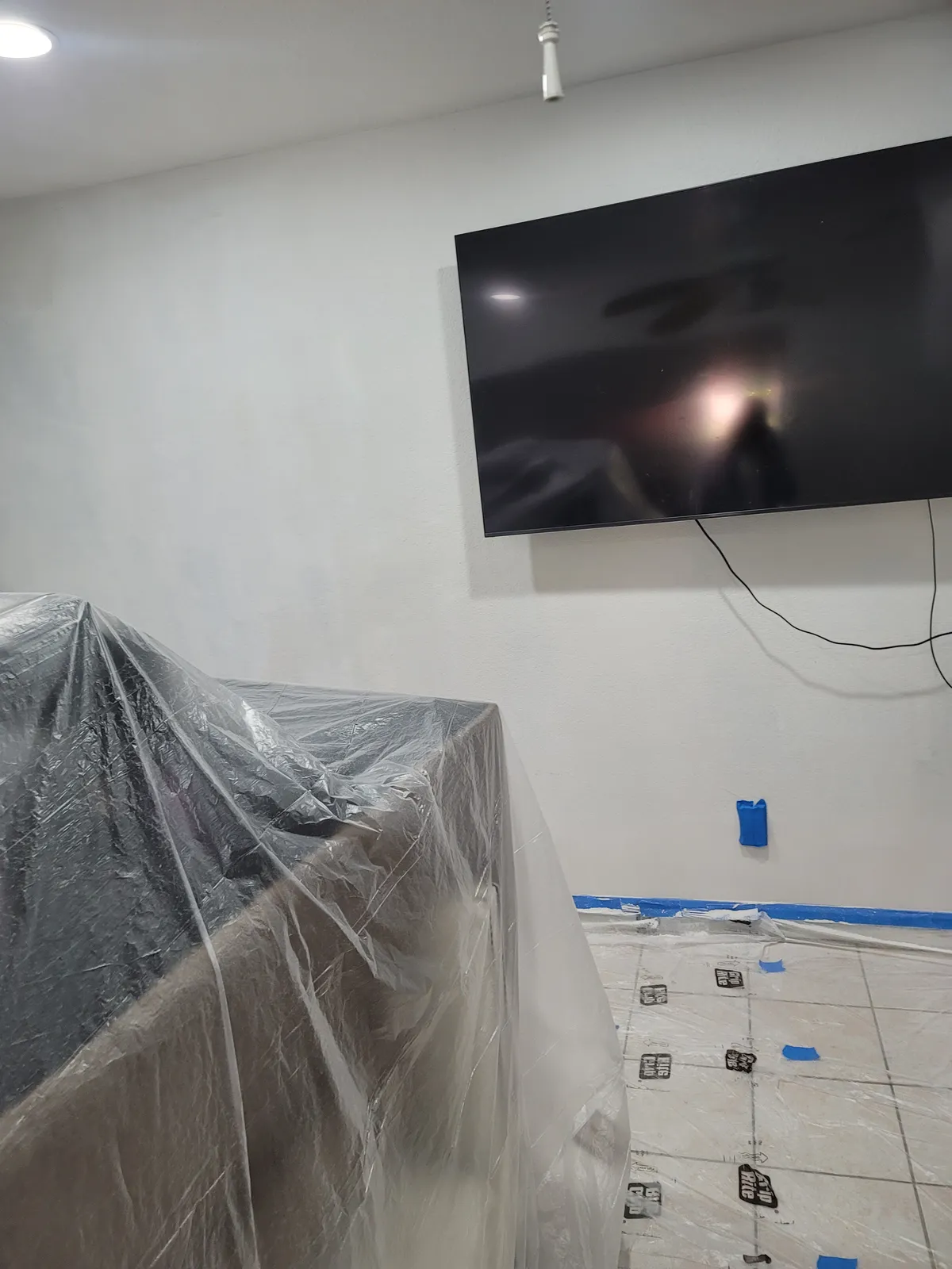 Interior room with furniture wrapped in plastic sheeting and the tiled floor papered and taped, beside a freshly painted wall with a wall-mounted television