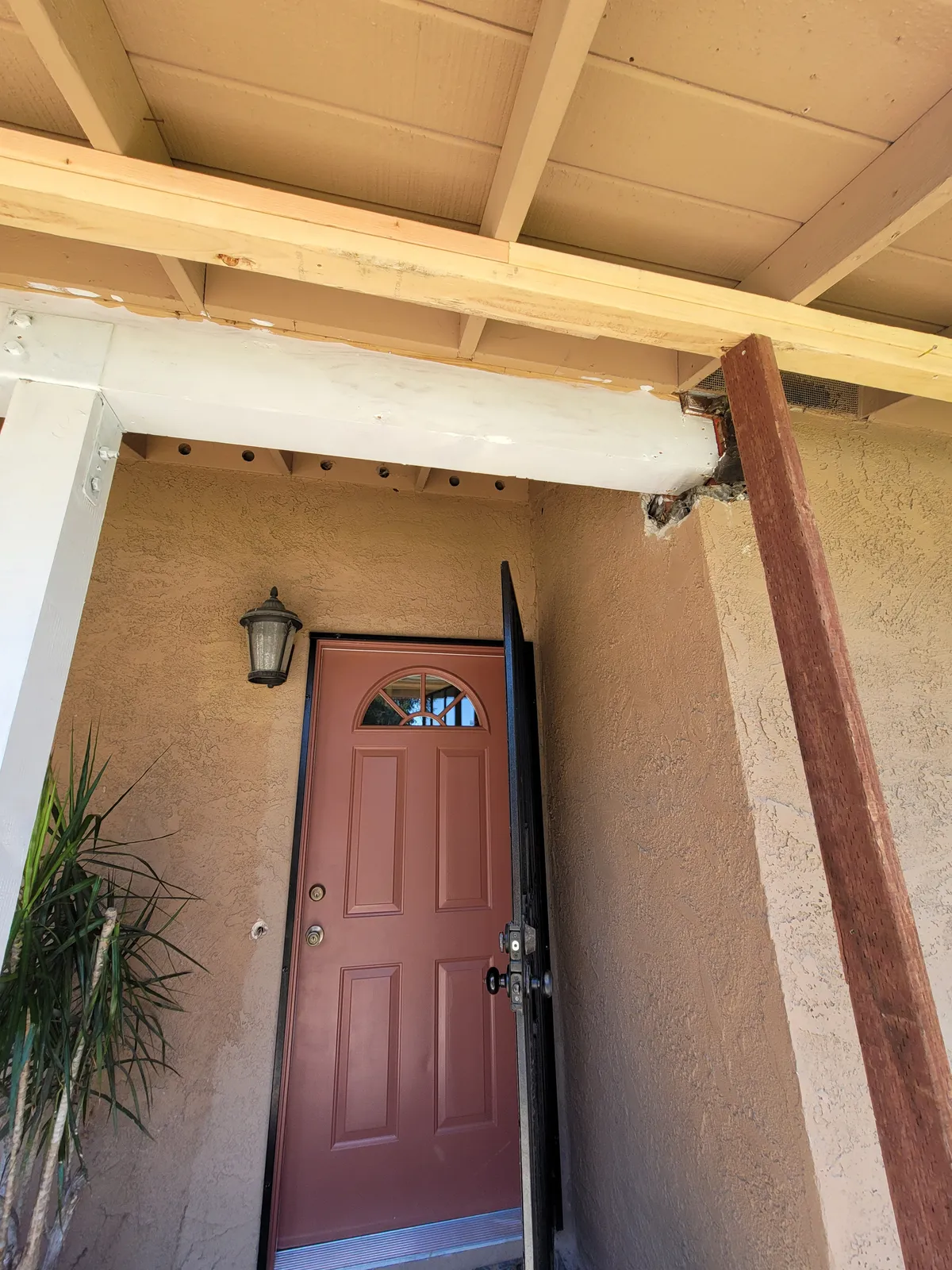 Entry porch shot from below, with a painted beam whose end is split and deteriorated where it meets the post, and a bare timber post beside a red front door