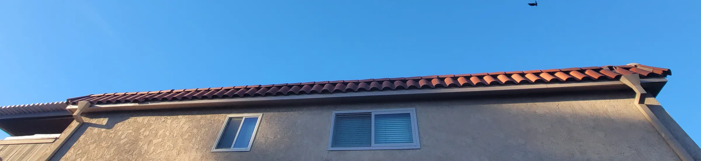 Long view along the roof line of a two-story stucco building, with the tile roof edge, a clear gutter run and a deep blue sky above