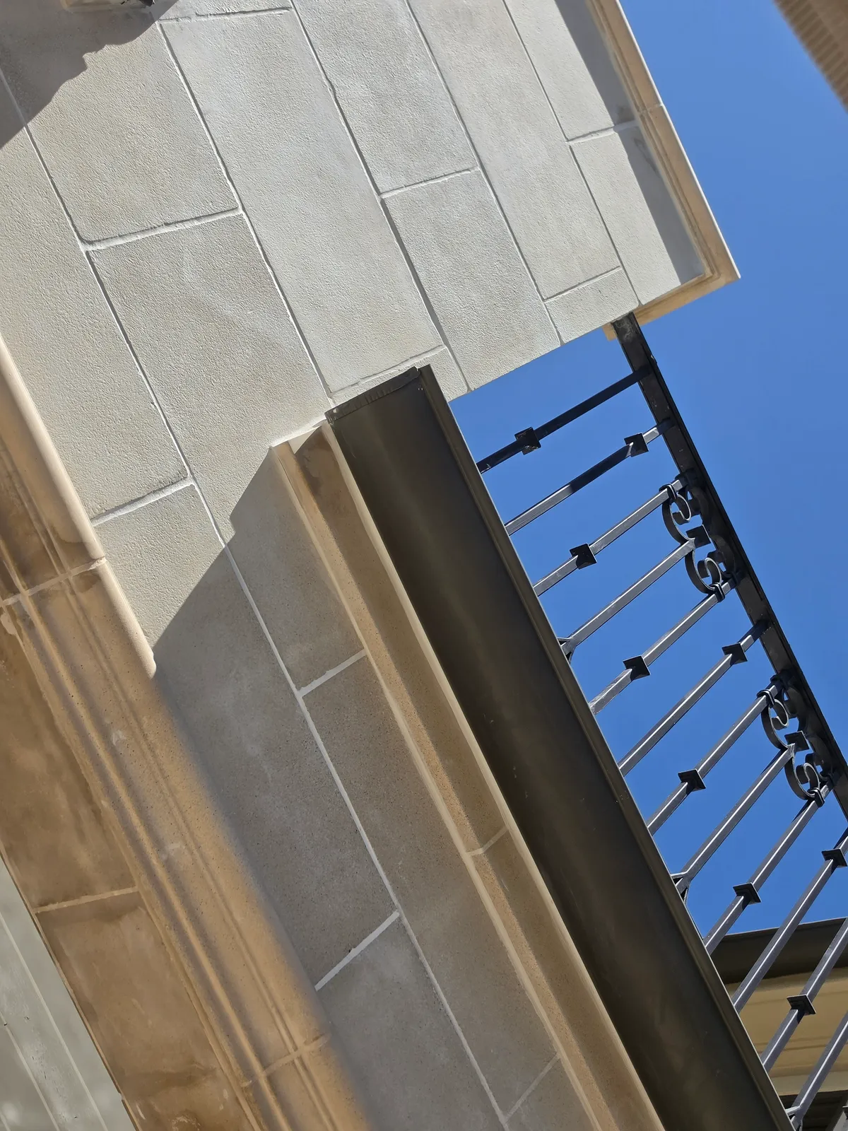 Looking up at pale stone-clad walls meeting a balcony soffit, with a black ornamental wrought iron railing against a deep blue sky