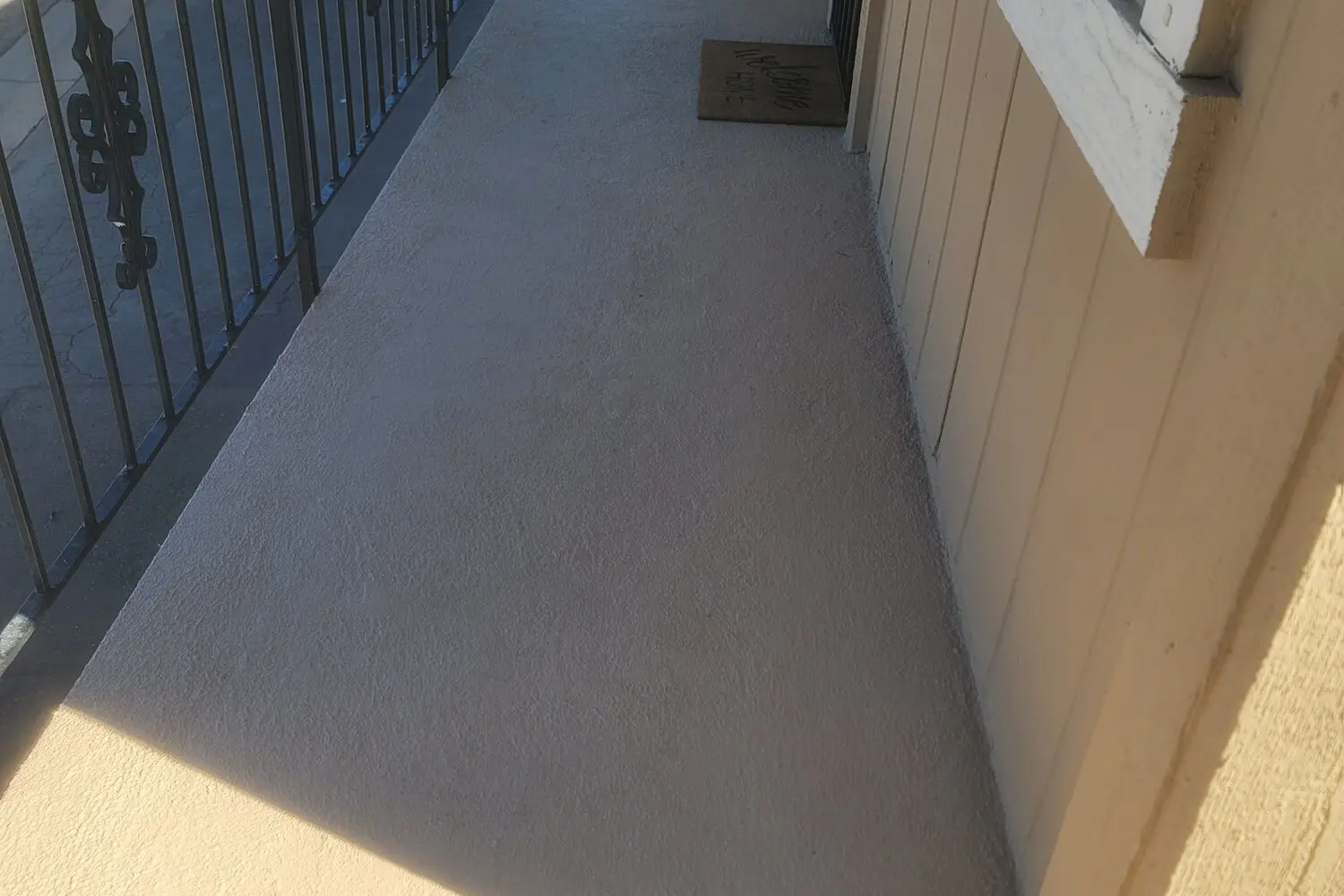 Freshly coated grey walkway deck at a condo building, with a black metal railing on the left and an entry mat by the door at the far end